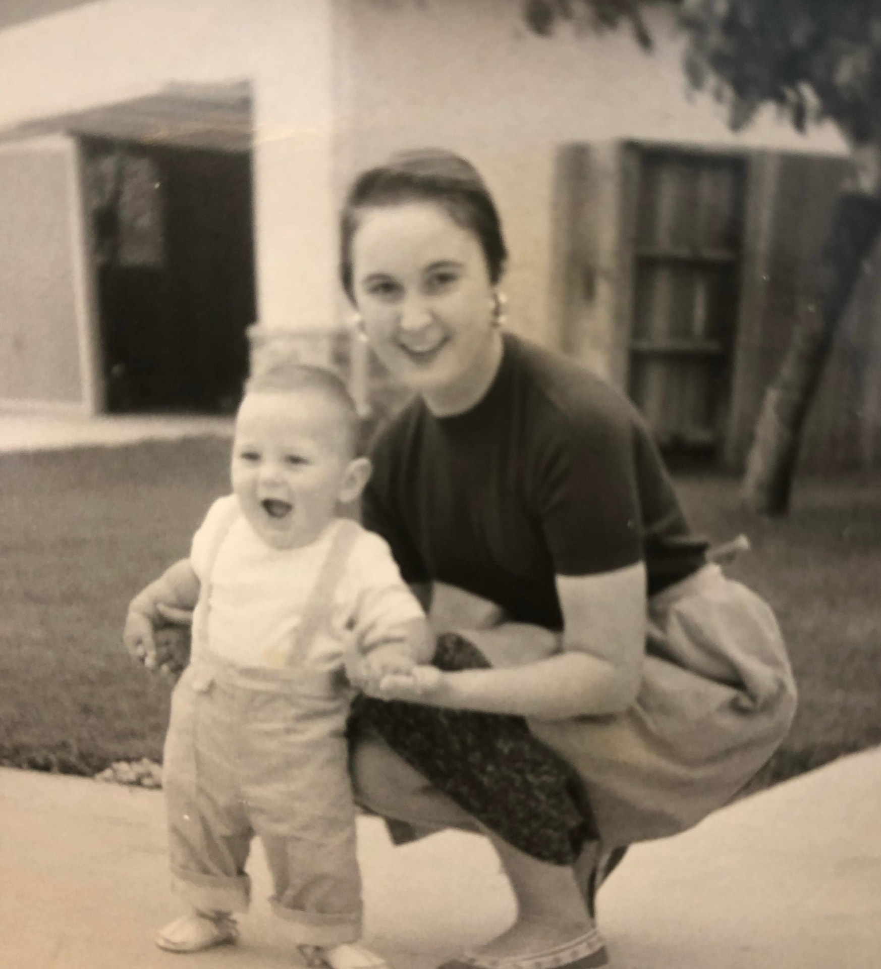 Faded black and white photograph of a young woman crouching on a driveway beside a laughing toddler in dungarees